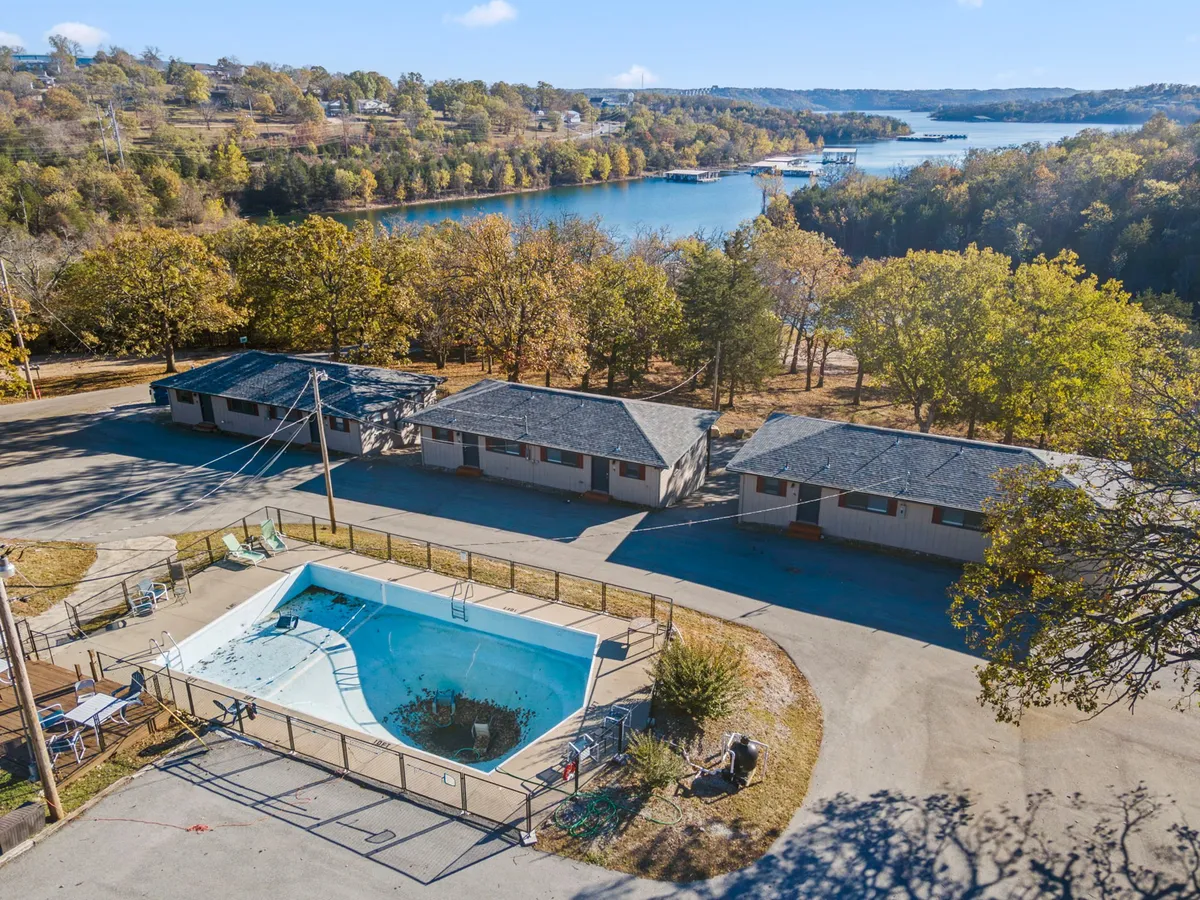 A sparkling outdoor swimming pool sits nestled amidst lush greenery with a clear view of the adjacent lake. The pool area is surrounded by trees and motel-style buildings, creating a relaxed and inviting resort atmosphere. Sun loungers line the water's edge, and the lake glistens in the background, suggesting opportunities for both swimming and waterfront relaxation.