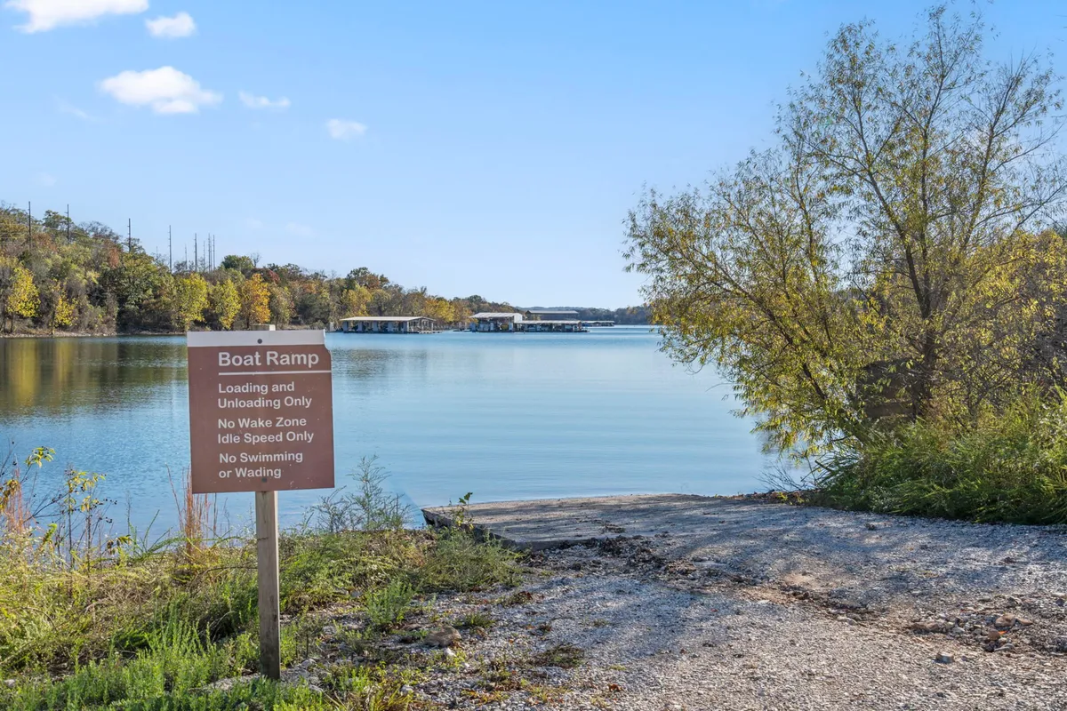 A serene lakeside view showcasing a boat ramp leading into calm waters, framed by lush trees and subtle signage. The scene hints at the ease of launching a boat and enjoying peaceful moments on the lake, with the sun low in the sky adding a gentle warmth to the environment.
