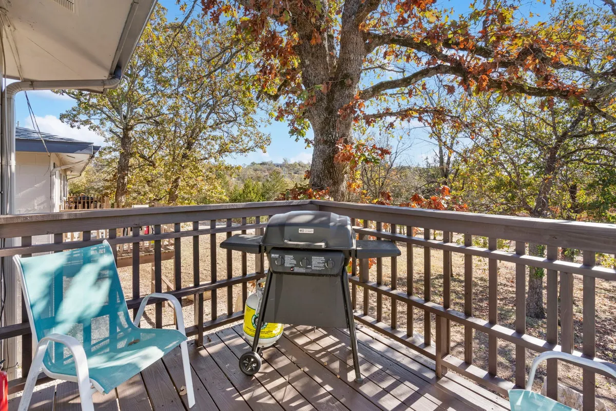 An inviting outdoor patio area with a grill, multiple outdoor chairs, and a canopy of trees overhead. The scene suggests gatherings with food, relaxation, and socializing in a casual, nature-connected setting.