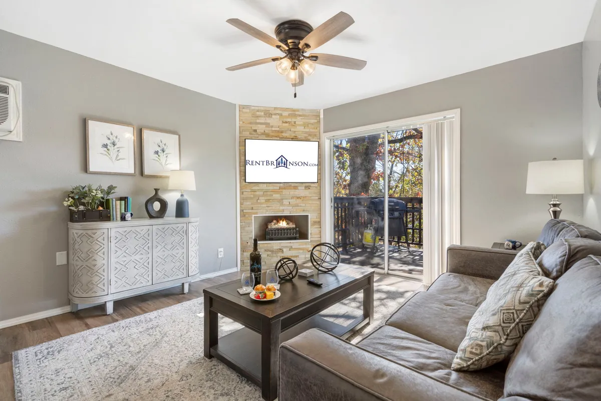 A stylish living space anchored by a modern fireplace, with a comfortable sofa, coffee table set with wine and snacks, and large windows opening out to the private deck. The room is bathed in natural light, and the view through the windows hints at the scenic outdoors. The setup is ideal for both relaxing evenings and social gatherings.