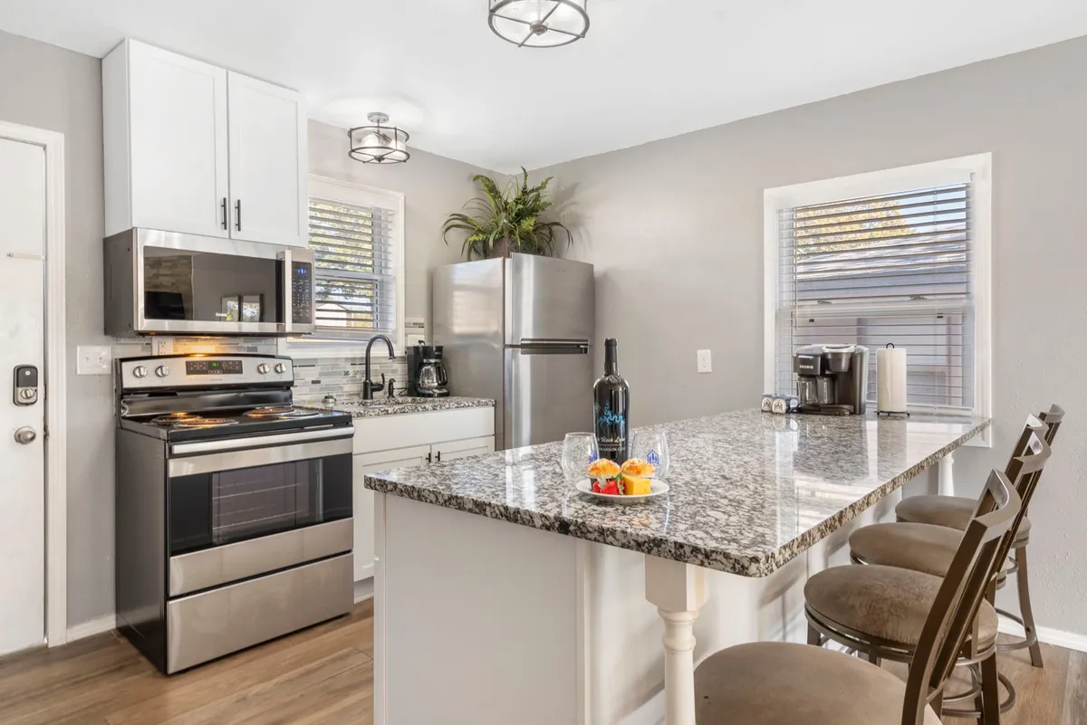 This image highlights a bright, modern kitchen area with granite countertops, stainless steel appliances, and bar stools. Decorative elements like a fruit bowl and window plants add warmth to the space. The scene is set for cooking, entertaining, or enjoying a casual breakfast.