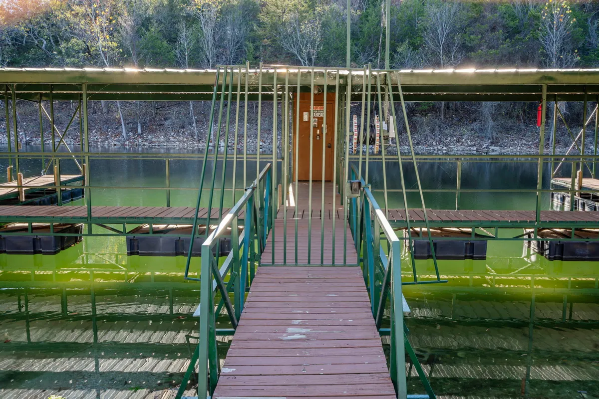 A peaceful scene featuring a wooden walkway leading to a boathouse surrounded by towering trees. The calm water reflects the sky and trees, evoking a tranquil lakeside atmosphere. People can be seen fishing from the dock, while a small boat floats nearby, highlighting the property's direct access to Table Rock Lake.