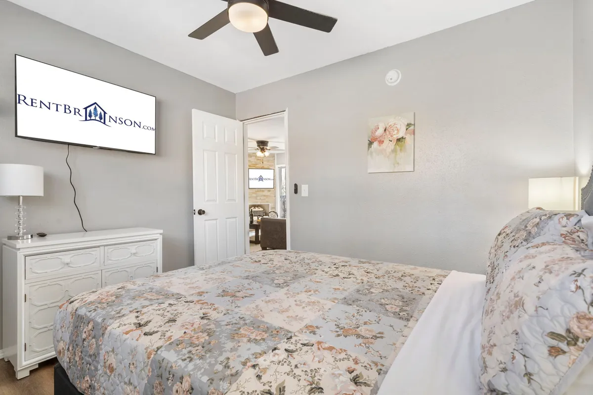 A cozy queen bedroom featuring a bed with floral bedding, a nightstand with a lamp, a mounted TV, and decorative wall art. The room exudes a warm and inviting atmosphere perfect for relaxation after a long day.
