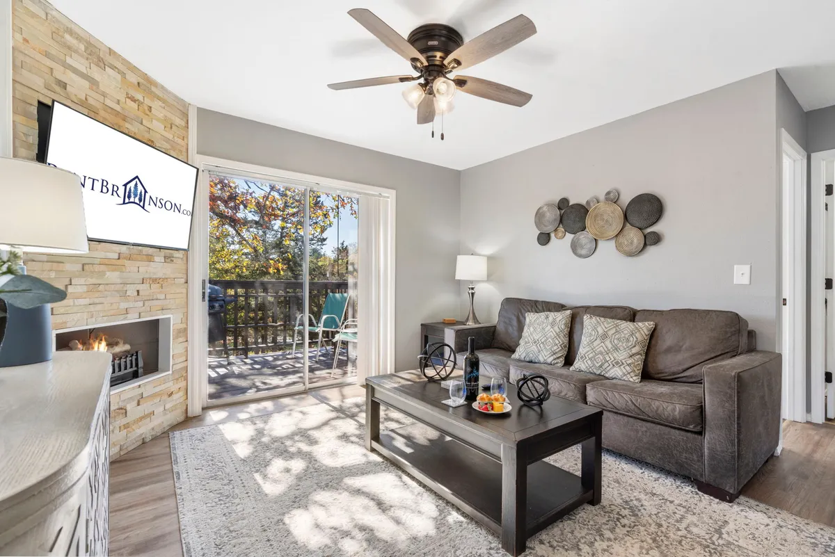 A welcoming living area featuring a comfortable sofa, flat-screen TV, sliding glass door leading to a private deck, decorative wall plates, and a coffee table topped with snacks. Sunlight streams in from the large windows, brightening the space and creating a cozy atmosphere for gathering, watching TV, or relaxing with friends and family.