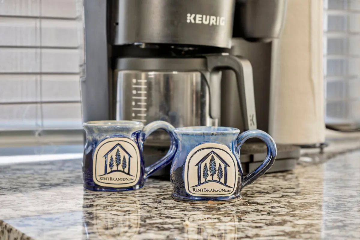 A close-up of two ceramic mugs beside a Keurig coffee maker on a granite countertop, set in a cozy corner. The scene suggests the simple pleasure of enjoying morning coffee with loved ones, perhaps while taking in lake views from nearby windows or the deck.