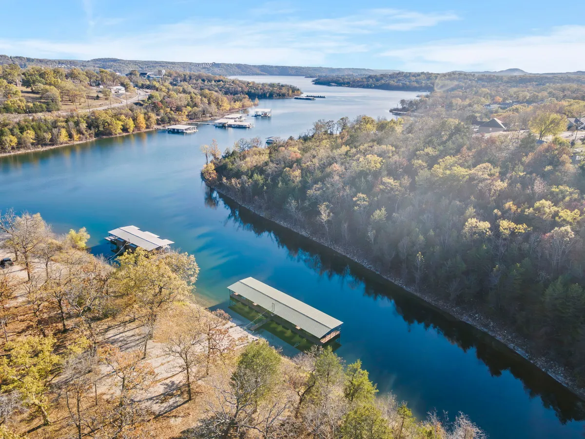 A wide scenic view highlights the calm water surface of the lake, boathouses lining the shore, and rolling, tree-covered hills in the background. The peaceful setting evokes the area’s natural beauty and opportunities for boating, fishing, picnicking, and hiking.