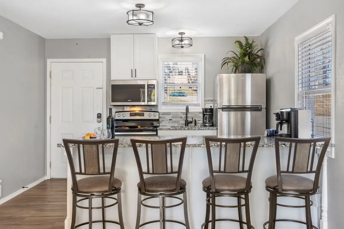 A slightly different angle of the kitchen, emphasizing the granite countertop, stainless steel appliances, and bar stools, with a kitchen window that brings in natural light. The setup encourages socializing and shared meal preparation.
