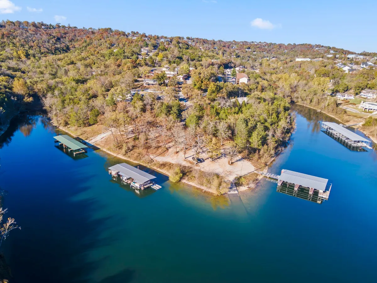 A tranquil view of calm blue lake waters bordered by docks, tree-covered hills, and residential homes. The setting is serene, with hints of outdoor activities like boating and fishing possible in the scene.